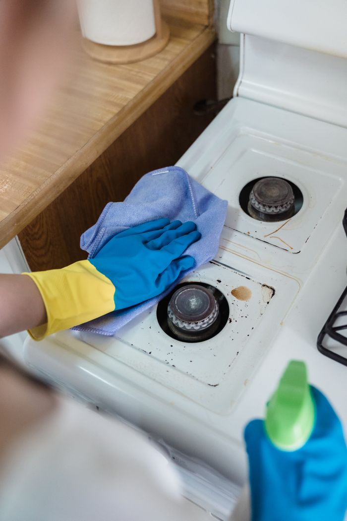 who-we-are A person wearing rubber gloves cleaning a dirty gas stove with a cloth and spray bottle.