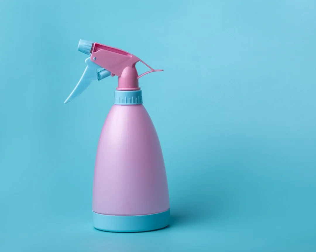 A woman wearing a plaid shirt cleans a kitchen counter using a spray bottle and cloth.