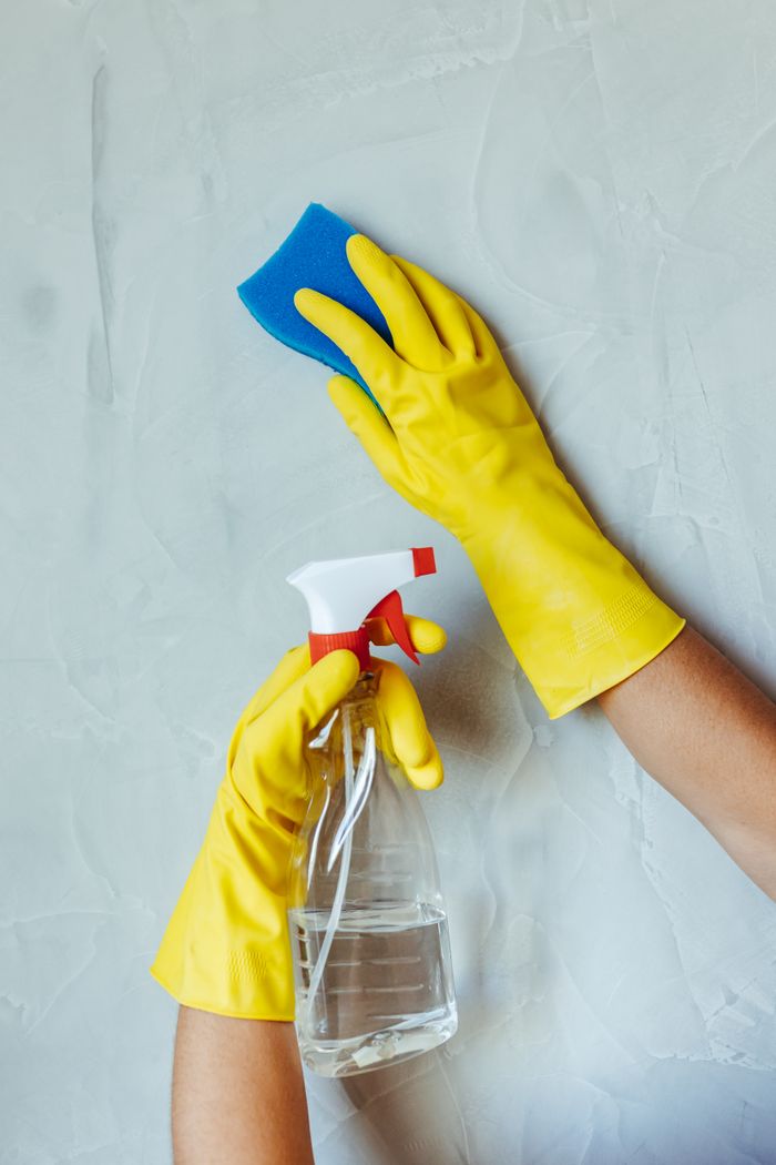 Hands in yellow gloves wiping a wall with a sponge and spray bottle.